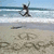 A beach scene with a person in dark clothing holding an object, sand drawings including peace signs and circles, ocean waves, and blue sky.