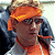 Pixelated image of a person wearing an orange head covering and dark clothing, holding an object near their face.