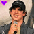 A person wearing a black cap and dark jacket holds a microphone, with a purple heart symbol in the top-left corner.