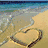Beach scene with heart-shaped sand drawing, ocean waves, and blue sky background.