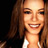 Pixelated close-up of a smiling person with long brown hair and light skin, wearing a white top against dark background.