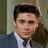 Pixelated static image of a person with dark hair, wearing a dark suit jacket over a white shirt and dark tie, against a muted background.