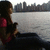 A pixelated static image shows a person with dark hair and red clothing seated near water, facing right toward a city skyline.