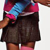 A cropped view of a person wearing a blue and pink striped long-sleeve shirt, a dark pleated skirt, and pink socks, with arms extended.