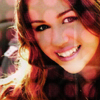 A close-up portrait of a person with long wavy brown hair, wearing a dark patterned top, smiling broadly with visible teeth.