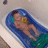 A child lies in a blue bathtub with green base, holding a yellow ring toy while water splashes around them.