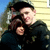 Two people embrace outdoors; the person in front wears a dark cap and dark hoodie with light patterns, the person behind has brown hair and a dark top, against a light-colored building with blue sky.