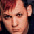 Pixelated close-up of a person with short red hair and light skin, wearing dark clothing against a dark background.