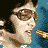 Pixelated image of a person with dark hair and sunglasses holding a microphone, set against blue and yellow backgrounds.