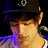 Pixelated close-up of a person wearing a dark hat with a light emblem, brown hair, and a purple garment against a dark background.