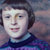 Pixelated static image of a person with short dark hair wearing a dark blue top against a blurred background with purple and blue tones.