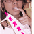 A person wearing a white sash with pink X symbols and a pink 'S' on the left side, holding a finger near their mouth, with a light brown background.