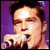 Pixelated static image of a person with dark hair against purple background, holding microphone near mouth.