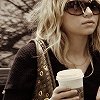 A person with blonde hair holds a white disposable coffee cup, wearing dark sunglasses and a black top with a patterned strap over the shoulder.