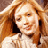 Pixelated static image of a person with long light brown hair wearing a white top against a blurred background.