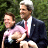 Static pixelated image of two men in formal attire with one holding a child, set against green foliage background.