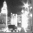 Static grayscale image of a stylized cityscape with tall buildings and bright light sources against dark background.