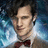 A person with dark tousled hair wears a brown suit jacket, white shirt, and red bow tie against a blue swirling background.