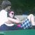 Two people in a boat, one person in white shirt and dark pants holding another person in blue checkered shorts and pink top, both seated on green surface with dark background.