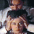 Two figures in close physical proximity, one positioned above the other, with visible facial features and dark hair against a light background.