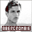 Pixelated grayscale portrait of a person with dark hair and red highlights, wearing a dark shirt, with text reading 'ABERCROMBIE' in white block letters on a dark brown bar below.