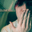 Pixelated image of a person with light skin covering their face with both hands, wearing a white top against a green background.
