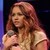 A pixelated image of a person with long brown hair wearing a light-colored sleeveless top, holding a microphone against a dark background.