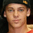 Pixelated close-up of a person wearing dark cap with small yellow and orange emblem, light skin tone, visible ear with earring.