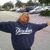A person with light brown hair stands outdoors in a parking lot wearing a dark blue hoodie, arms outstretched.
