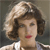 Pixelated portrait of a person with light brown hair styled in a bouffant, wearing red lipstick and a white garment with a dark collar.