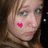 Pixelated close-up of a person's face with a bright pink heart symbol on the left cheek, light brown hair, and blue eyes, set against a dark background with small colored squares.