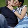 Two people kiss on a grassy background; one wears a checkered shirt and blue jeans, the other a dark jacket and light pants.
