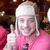 A person with light-colored hair wears a white head covering and pink clothing, making a peace sign with their right hand against a dark background.