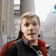 A person in a black jacket with red collar and dark shirt stands against a blurred urban background, displaying sequential facial expressions from neutral to smiling.