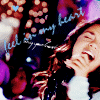 A person with long brown hair in a light blue top, hand near mouth, against a dark background with purple and orange bokeh lights and text reading "feel in my heart" with a small blue heart symbol.
