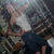 A person in a dark blue shirt makes a peace sign with their right hand against a blurred background of indistinct shapes and colors.