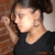 A woman with dark hair in a bun wears a black top and large hoop earrings, shown in three sequential poses against a brick wall background.