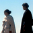 Two pixelated figures standing side-by-side against a light blue background, one in light clothing and the other in dark clothing.