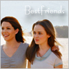 Two women stand side-by-side outdoors with a body of water and distant land in the background, wearing casual tops under soft lighting.