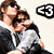 Two people in formal attire embrace; one wears a black jacket over white shirt, the other has dark hair and sunglasses. A stylized 'E3' logo appears to their right.