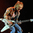 Pixelated image of a person with light brown hair playing an electric guitar, wearing blue jeans and a sleeveless top against dark background.