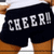 A person wearing black shorts with white text 'CHEER!' on the back, viewed from behind with a white top and brown pants visible.