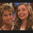Two people with light brown hair stand side-by-side, smiling at the camera against a blurred background.