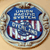 Circular badge with blue top section, red and white striped middle, and blue bottom section, bordered by brown and white rings on a beige background.