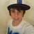 Pixelated image of a person wearing a black hat with a blue top, white shirt, and blue pants, smiling against a light beige background.
