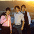 Three figures stand in a against a warm-toned background; the leftmost figure wears pink, middle white, right dark vest over light shirt