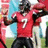 Pixelated image of a person in a red jersey with white number 7, black shorts, and dark gloves, standing with arms raised against a blurred background.