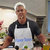 Pixelated image of a person wearing a white apron over dark long sleeves, standing in a kitchen with blurred background elements.