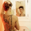 A woman with short dark hair styled in an updo adjusts her hair while facing a mirror, wearing a backless dress with crisscross straps.