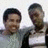 Two people in a static pose with one person smiling broadly and the other looking down, both in light-colored shirts with dark outer layers against a plain background.